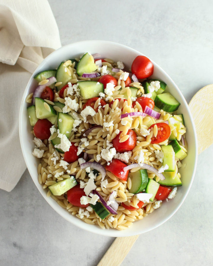 greek orzo pasta salad in a bowl with a towel and wooden spoon in the background