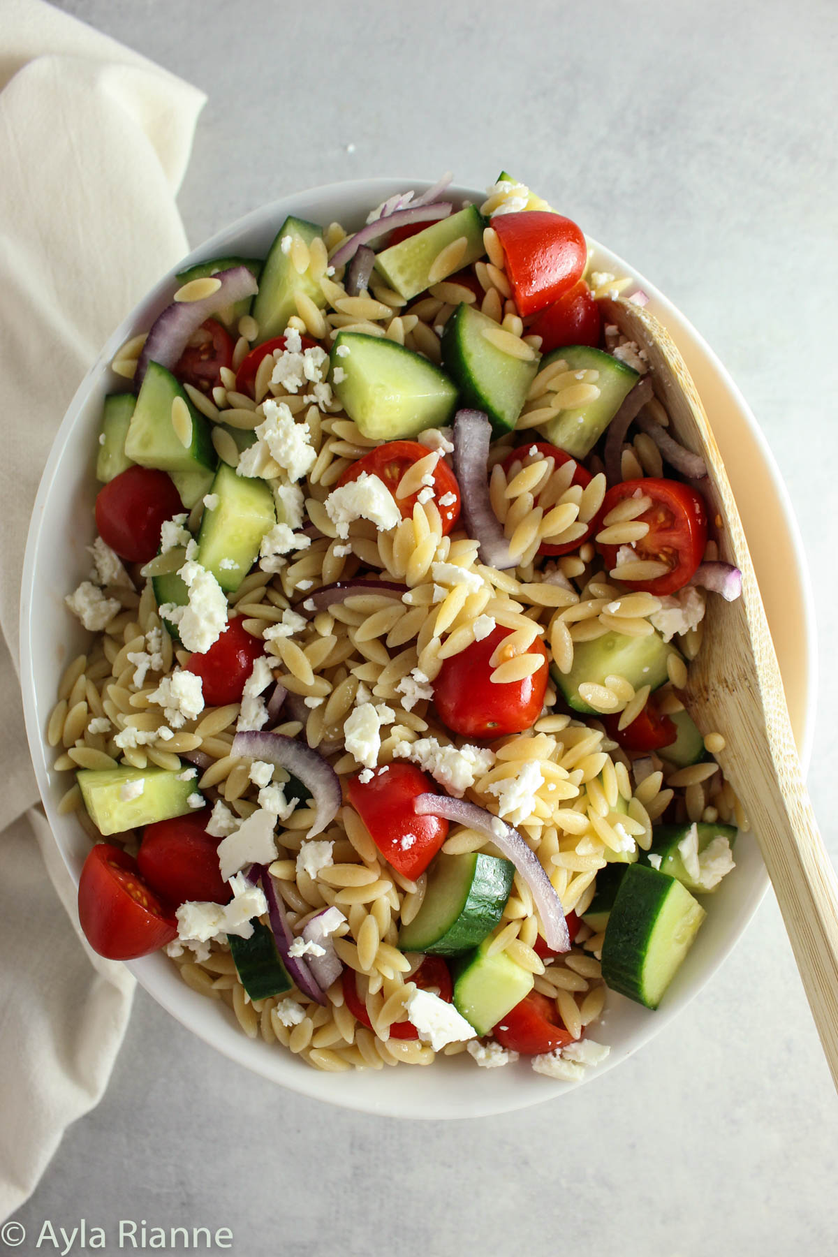 greek orzo pasta salad in a bowl with a wooden spoon in the bowl and a towel in the background